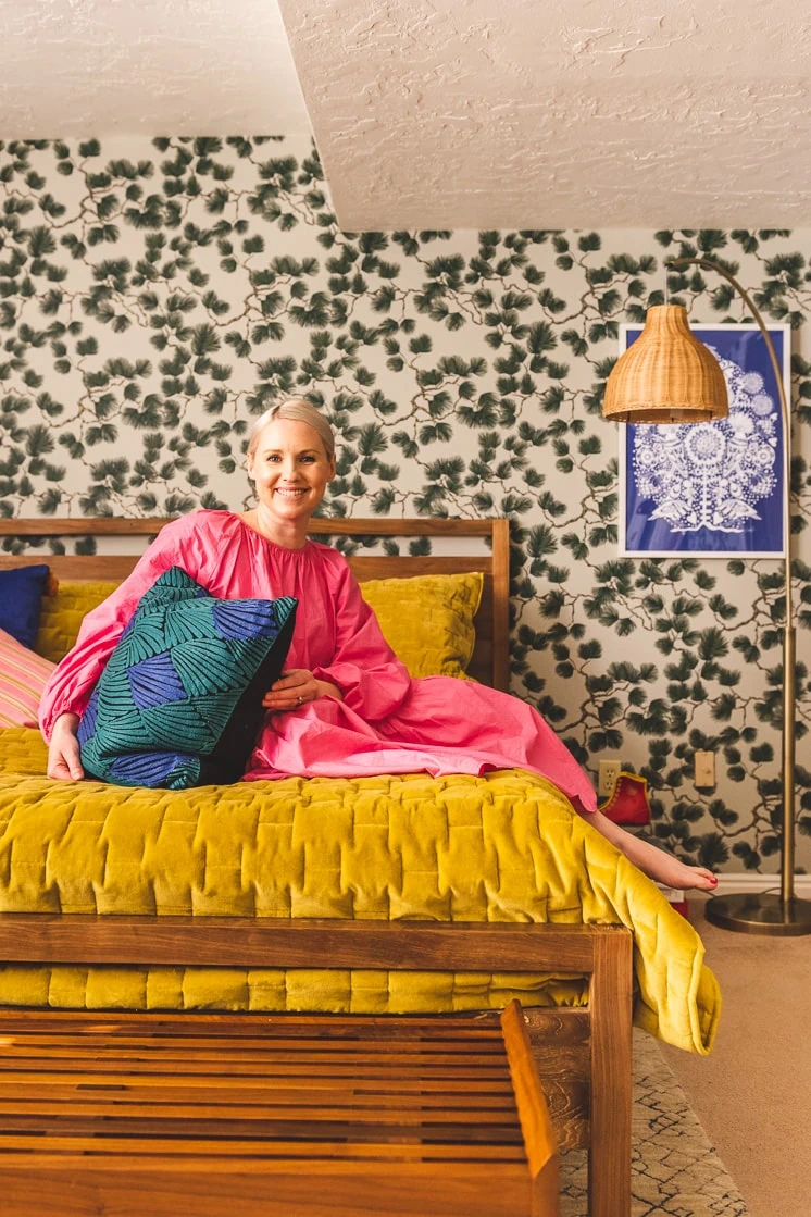 Brittany sits on bed in a room with pine-themed wallpaper. She's wearing a pink dress and holding a dark blue and green pillow, the bed is warm wood with a mustard duvet, and there's a wicker lamp in the corner. There's also a blue art print on the wall.